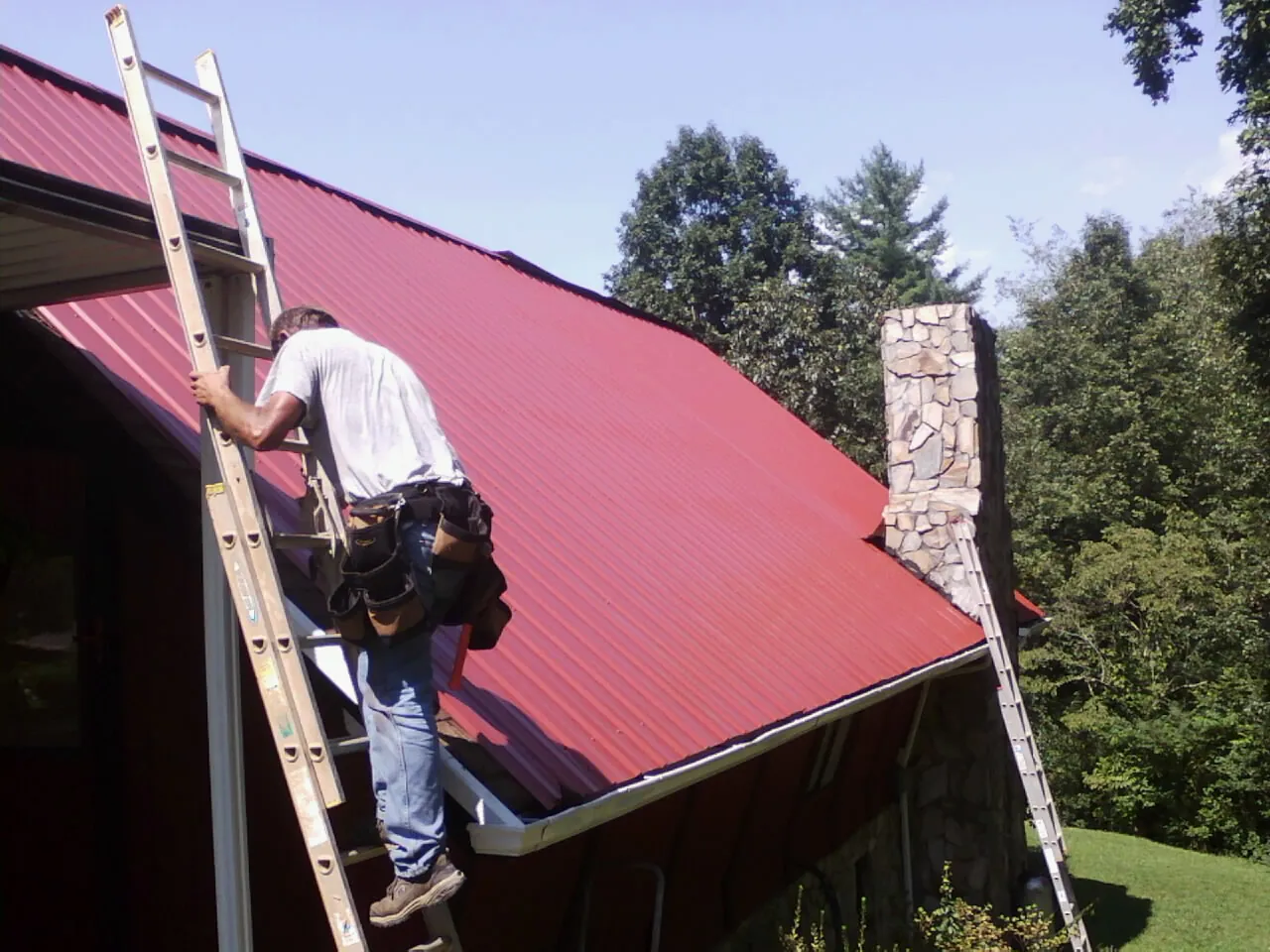 Roofer on a ladder beside a red metal roof and stone chimney — Soffit & Fascia Repair by Roofing Brothers in Island Heights